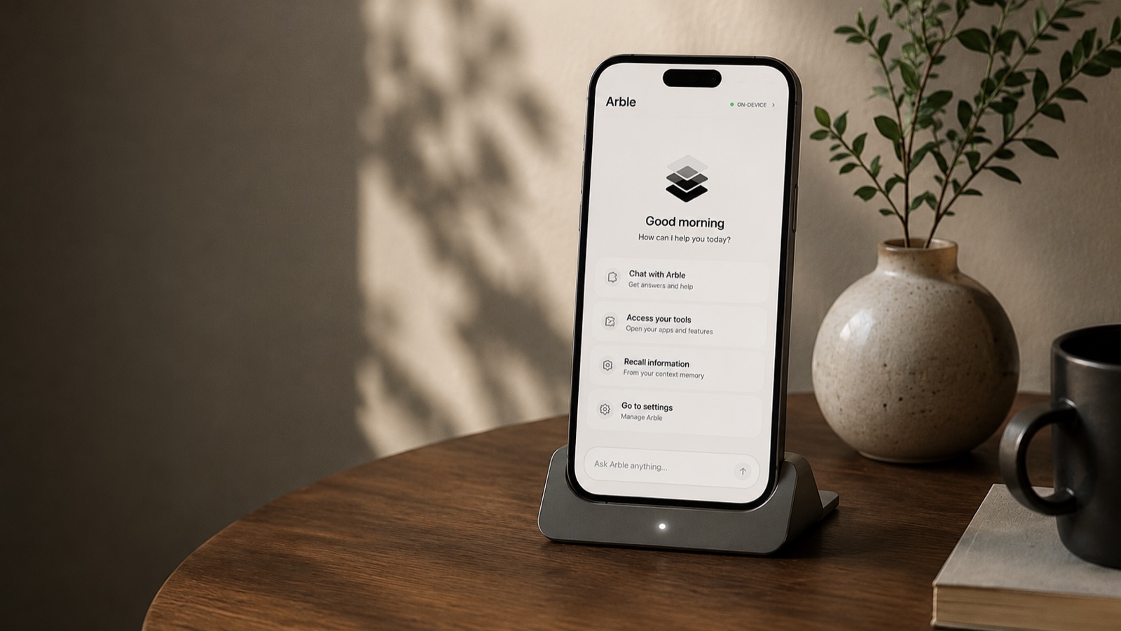 An iPhone standing in a dock on a wooden side table, showing Arble's home screen — a Good morning greeting, an ON-DEVICE badge, and shortcuts to chat, tools, context memory and settings — beside a potted plant and a mug.